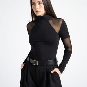 Op de afbeelding: Een zwart shirt met lange mouwen en een opstaande kraag, voorzien van transparante mesh-panelen op de schouders en bovenarmen, gecombineerd met een zwarte broek met hoge taille en een zwarte leren riem met zilverkleurige gesp.