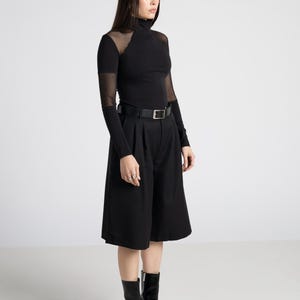 May include: Black ensemble featuring a long-sleeved turtleneck top with sheer mesh accents on the shoulders and sleeves, paired with high-waisted culottes and a black belt. The outfit is completed with black heeled boots. The overall style is modern and minimalist.