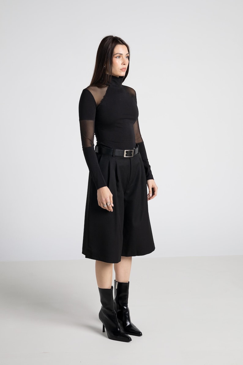 May include: Black ensemble featuring a long-sleeved turtleneck top with sheer mesh accents on the shoulders and sleeves, paired with high-waisted culottes and a black belt. The outfit is completed with black heeled boots. The overall style is modern and minimalist.