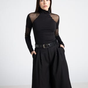 May include: Black turtleneck top with sheer mesh shoulder and sleeve accents, paired with high-waisted black shorts and a black belt. The outfit is a monochromatic ensemble, creating a sleek and modern look.
