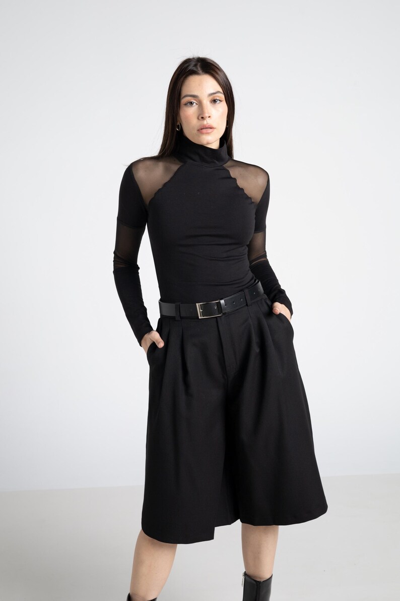 May include: Black turtleneck top with sheer mesh shoulder and sleeve accents, paired with high-waisted black shorts and a black belt. The outfit is a monochromatic ensemble, creating a sleek and modern look.