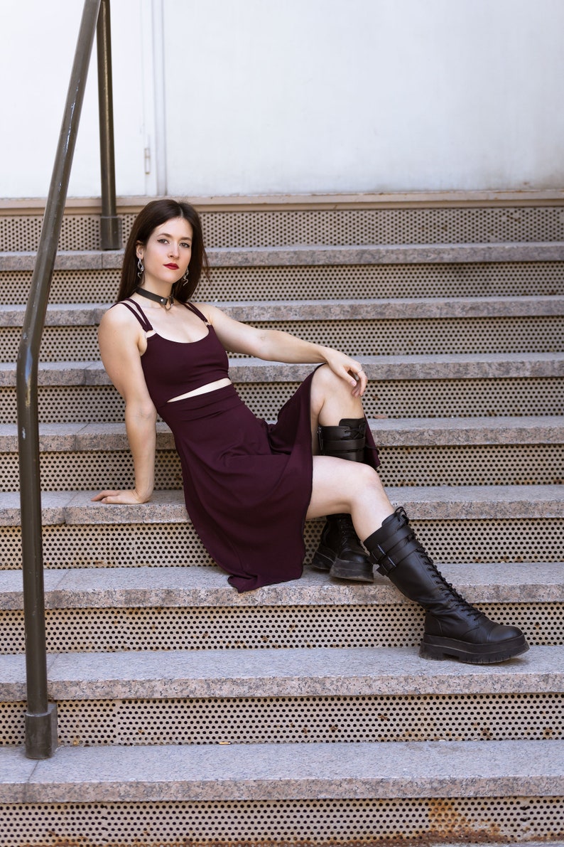 May include: A woman wearing a burgundy dress and black boots sits on a set of concrete stairs with a metal railing.