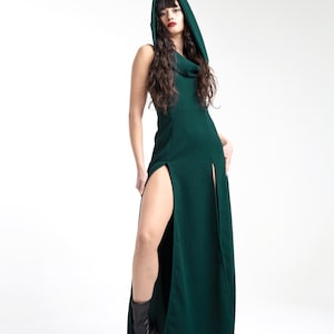 May include: A woman wearing a long, dark green dress with a cowl neck and a hood. The dress has a high slit on each side. She is wearing black boots.