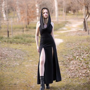 Puede incluir: Una mujer con un vestido negro maxi con capucha y una abertura en el muslo. El vestido tiene un cuello de pico y está hecho de una tela suave y fluida. Está de pie en un área boscosa con un camino frente a ella.