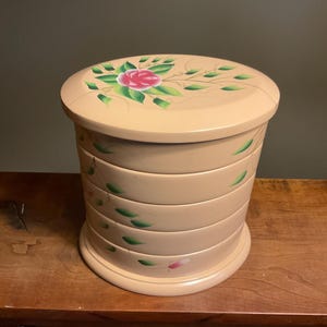 May include: A beige, round, multi-tiered jewelry box with a painted floral design. The lid features a pink rose with green leaves and vines.