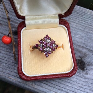 May include: A red and white gemstone ring in a red velvet ring box. The ring features a cluster of red gemstones surrounded by smaller white gemstones.