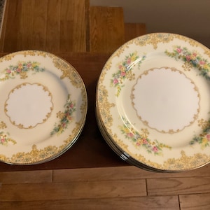 May include: A set of cream-colored porcelain plates with a floral and gold filigree design. The plates have a scalloped edge and a white center. The floral design includes pink, blue, and yellow flowers.