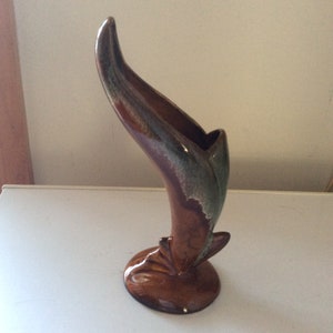 May include: A brown and green ceramic vase shaped like a curved horn with a wide opening at the top. The vase has a brown base.