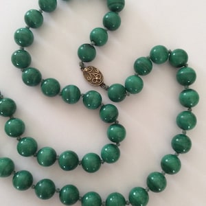 May include: A long necklace made of green malachite beads with a gold clasp. The beads are round and smooth, and the clasp is ornate.