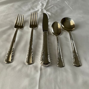 May include: A set of five silver-plated flatware pieces, including two forks, a knife, and two spoons. The flatware has an ornate design and is arranged on a white tablecloth.