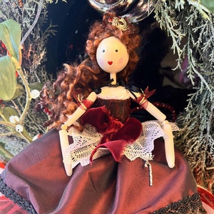 May include: A handcrafted doll with a wooden head, curly auburn hair, and a red heart-shaped mouth. The doll wears a burgundy dress with a white lace trim, a black bodice, and a red velvet bow. The doll is decorated with a small gold crown.