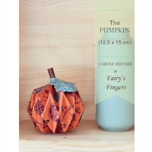 Puede incluir: Una calabaza de papel plegada con un patrón floral, que mide 12,5 cm por 15 cm. La calabaza es naranja con un tallo y una hoja verdes. El texto "The PUMPKIN (12,5 x 15 cm) CAROLE MEUNIER is Fairy's Fingers" está sobre un fondo azul claro.