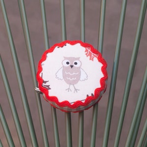 Peut inclure: Un ruban à mesurer en tissu rouge et blanc avec un motif de hibou. Le hibou est brun avec des yeux blancs et un ventre blanc. Le ruban est sur une chaise verte.