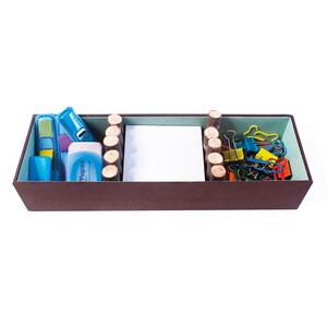 May include: A brown rectangular box with a light green interior. The box is divided into three sections. The left section contains blue and yellow office supplies, the middle section contains a white notecard, and the right section contains a variety of colorful paper clips.