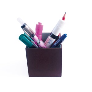 May include: A brown pencil holder with a pink highlighter, a green marker, a blue pen, a silver pen, and a red pencil inside.