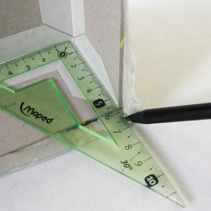 May include: A green protractor with a black pencil marking a line on a piece of white fabric. The protractor has markings in centimeters and inches. The protractor has the word 'Maped' printed on it. The protractor is measuring a 30 degree angle.
