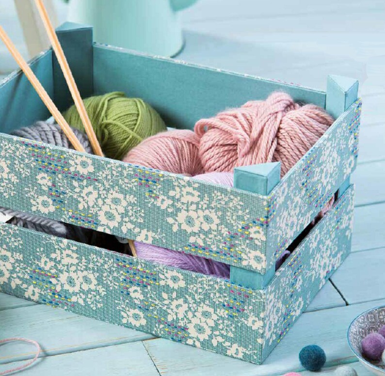 May include: A blue wooden crate with a floral pattern lining the inside. The crate is filled with balls of yarn in various shades of pink and green.