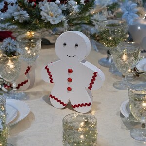 May include: White gingerbread man cutout with red trim and red button accents. The gingerbread man is on a white tablecloth with clear glass candle holders and white plates.