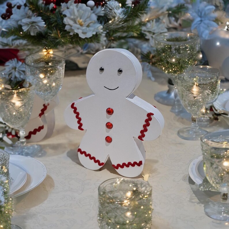 May include: White gingerbread man cutout with red trim and red button accents. The gingerbread man is on a white tablecloth with clear glass candle holders and white plates.