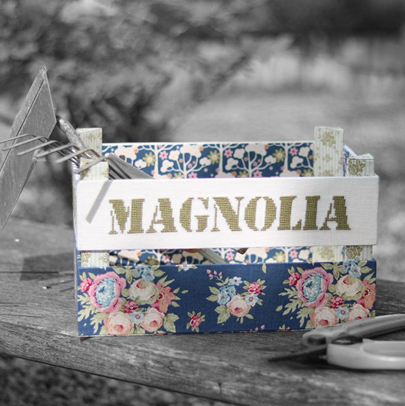 May include: A blue and white floral patterned box with a white label that says 'MAGNOLIA' in green letters. The box is sitting on a wooden surface.