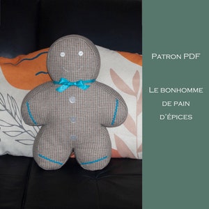Puede incluir: Un juguete de peluche de jengibre marrón con una pajarita azul y botones blancos. El juguete está sentado en un sofá con una almohada estampada detrás. El texto "PATRON PDF LE BONHOMME DE PAIN D'ÉPICES" es visible en el lado derecho de la imagen.