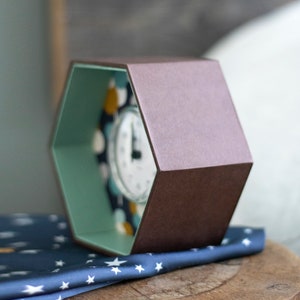 May include: A hexagonal box with a brown exterior and a teal interior. The box is open and a clock with a white face and black hands is visible inside. The box is sitting on a blue fabric with white stars.