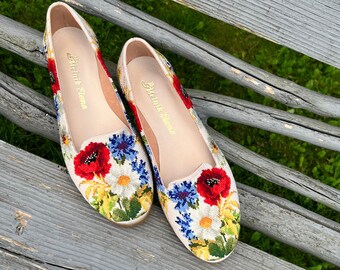 May include: A pair of floral embroidered flats with a cream-colored base. The flats feature a colorful design of red poppies, blue cornflowers, and yellow and white daisies. The shoes are displayed on a weathered wooden bench.
