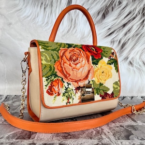 May include: A beige handbag with a floral embroidered design featuring roses, poppies, and other flowers. The bag has a brown leather handle and a gold chain strap.