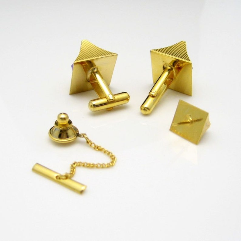 May include: A set of gold cufflinks and a tie tack with purple stones. The cufflinks are square with a pyramid shape on top. The tie tack is a pyramid shape with a small chain.