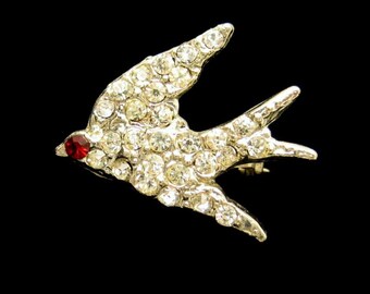 May include: A silver-coloured bird brooch, encrusted with clear rhinestones. The bird has a red rhinestone eye, set against a black background. This vintage-style brooch is an elegant accessory.