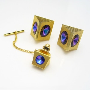 May include: A set of three gold-tone tie tacks with iridescent blue and purple stones. The tie tacks are shaped like cubes with a faceted stone on each side.