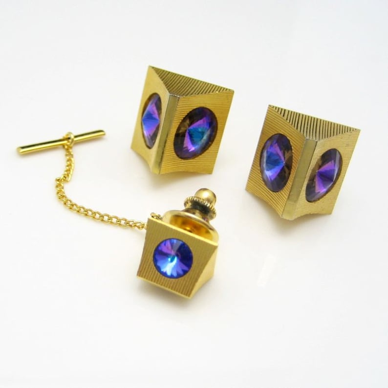 May include: A set of three gold-tone tie tacks with iridescent blue and purple stones. The tie tacks are shaped like cubes with a faceted stone on each side.