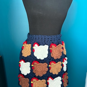 May include: A navy blue crochet skirt with a repeating pattern of brown, white, and tan chickens with red combs. The skirt is displayed on a black mannequin against a teal background. The crochet work is detailed and textured.