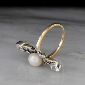 May include: A gold and silver ring featuring a white pearl and several small clear stones. The ring has a unique design with a curved gold band and a silver setting for the pearl and stones. The ring is set against a marble background.