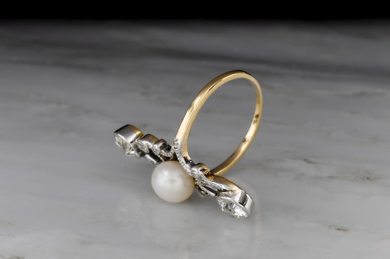May include: A gold and silver ring featuring a white pearl and several small clear stones. The ring has a unique design with a curved gold band and a silver setting for the pearl and stones. The ring is set against a marble background.