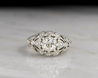 May include: A vintage engagement ring with a round brilliant cut diamond set in a filigree platinum setting. The ring is adorned with smaller diamonds around the center stone.