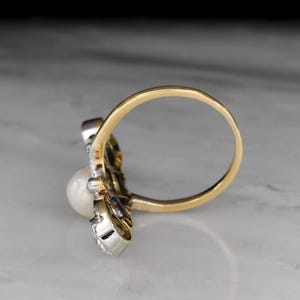 May include: A gold ring with a white pearl and small accent stones. The ring has a unique design with a gold band and a cluster of stones around the pearl. The ring is set against a marble surface.