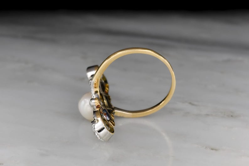 May include: A gold ring with a white pearl and small accent stones. The ring has a unique design with a gold band and a cluster of stones around the pearl. The ring is set against a marble surface.