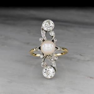 May include: An elegant ring featuring a central pearl and sparkling diamonds. The ring has a gold band and a silver-tone filigree design. The diamonds are round-cut and set in a decorative pattern. The ring is displayed on a marble surface.