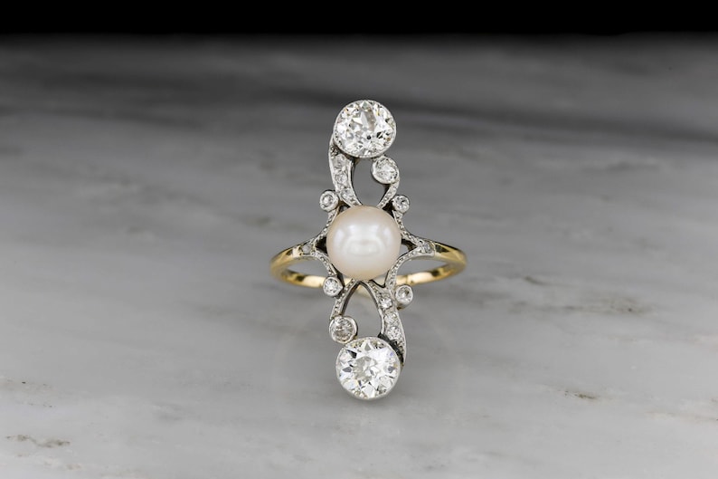 May include: An elegant ring featuring a central pearl and sparkling diamonds. The ring has a gold band and a silver-tone filigree design. The diamonds are round-cut and set in a decorative pattern. The ring is displayed on a marble surface.