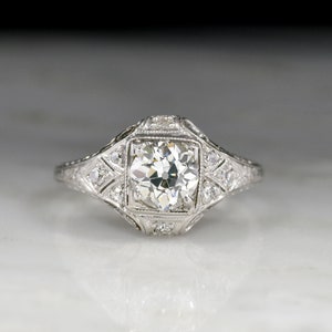 May include: A vintage engagement ring featuring a round brilliant cut diamond set in a platinum setting. The ring has a delicate filigree design and a halo of smaller diamonds.