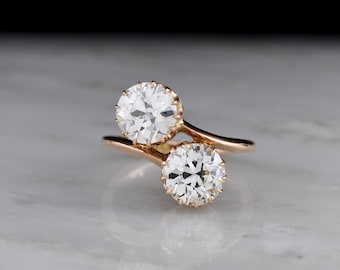 May include: A gold-toned ring featuring two large, round, clear gemstones. The gemstones are set in a swirling design, with the band curving around them. The ring is displayed on a reflective, light-colored surface.