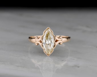 May include: A rose gold ring featuring a marquise-cut champagne-colored gemstone set in a decorative bezel with scrollwork details on the shoulders. The band is smooth and polished, showcasing a clear, elongated center stone with facets.