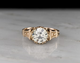 May include: A gold engagement ring with a large, round, clear diamond center stone. The ring has an ornate, filigree design on the band and around the diamond setting. The ring is set against a marble background.
