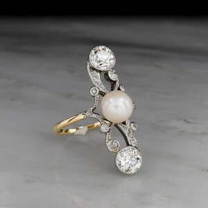 May include: An elegant ring featuring a large, lustrous pearl and sparkling diamonds. The ring has a unique, elongated design with a gold band and intricate silver detailing. The diamonds are set in a delicate, swirling pattern, adding to the ring's sophisticated appeal.