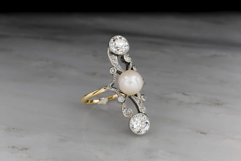 May include: An elegant ring featuring a large, lustrous pearl and sparkling diamonds. The ring has a unique, elongated design with a gold band and intricate silver detailing. The diamonds are set in a delicate, swirling pattern, adding to the ring's sophisticated appeal.