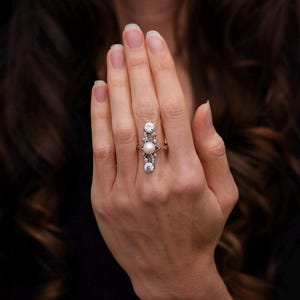 May include: A long, vertical ring featuring a central pearl, flanked by two larger, clear gemstones at the top and bottom. The ring is set in a silver or white metal, and is worn on a hand with a dark background.