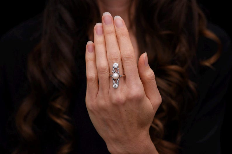 May include: A long, vertical ring featuring a central pearl, flanked by two larger, clear gemstones at the top and bottom. The ring is set in a silver or white metal, and is worn on a hand with a dark background.