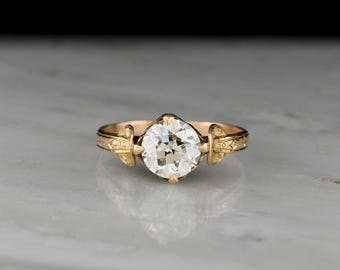May include: A gold ring featuring a single round, faceted clear stone set in the center. The band is crafted from yellow gold and displays intricate, engraved detailing on both sides of the central stone setting, which is held by four prongs.