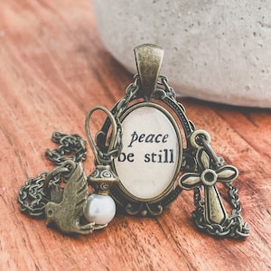 Può includere: Una collana dorata con un ciondolo che presenta un cammeo ovale bianco con le parole "peace be still". La collana include anche un piccolo ciondolo a forma di colomba e un ciondolo a forma di croce.